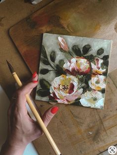 a woman holding a paintbrush next to a painting on a piece of paper with flowers painted on it