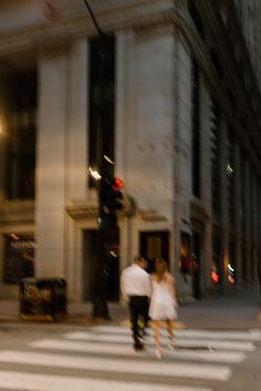 two people walking across a cross walk in front of a tall building at night time