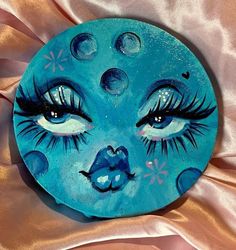 a painted plate with blue eyes and eyelashes on it's face, sitting on a pink satin surface