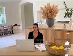 a woman sitting at a table with a laptop computer in front of her and a bowl of lemons on the counter