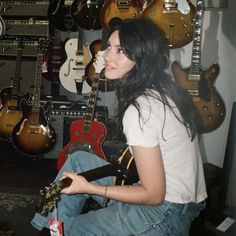 a woman sitting on the floor with her guitar in front of her and many guitars behind her