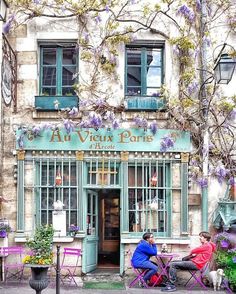 two people sitting at tables outside an old building with purple flowers on the front and green shutters