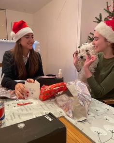 two women sitting at a table with presents and a small dog wearing a santa hat