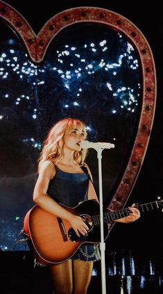 a woman holding a guitar while standing in front of a heart shaped stage with fireworks