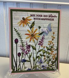 a card with flowers and butterflies on it, sitting on a clear stand in front of a white brick wall