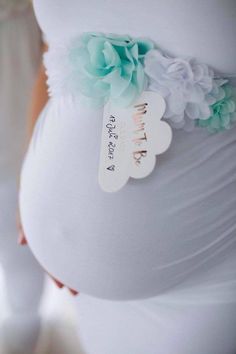 a pregnant woman wearing a white dress with flowers on the side and a tag attached to her belly