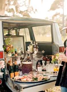 two people standing next to a car with food and drinks on the table in front of it