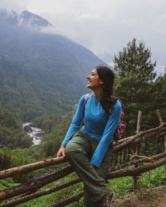 a woman standing on top of a wooden fence next to a lush green forest covered hillside