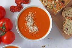 a bowl of tomato soup next to sliced bread and tomatoes