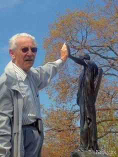 an older man standing next to a statue in front of a tree with leaves on it