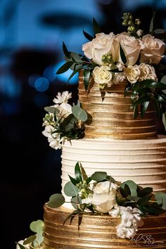 a three tiered cake with white flowers and greenery on the top is gold