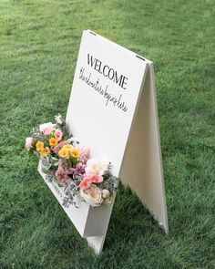 a welcome sign with flowers on it in the grass at a wedding ceremony or reception