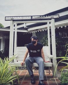 a man sitting on a bench in front of a house with plants and bushes around him