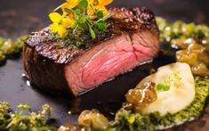 a piece of steak on a black plate with flowers and sauces around it, surrounded by green garnishes