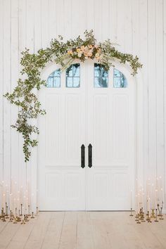 a white door with flowers and greenery on the top is surrounded by candles in front of it