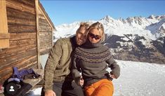 a man and woman sitting on top of a snow covered slope next to a wooden cabin