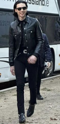 a man in black jacket and sunglasses walking next to a bus