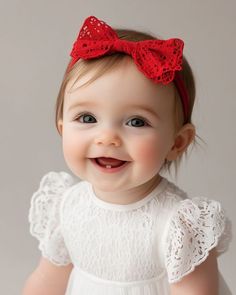 a smiling baby girl with a red bow on her head wearing a white lace dress