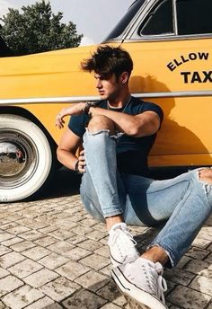 a man sitting on the ground in front of a yellow taxi car with his arms crossed