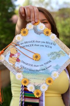 a woman holding up a winnie the pooh graduation cap with daisies on it