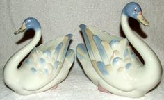 two ceramic birds sitting on top of a white blanket next to each other in the shape of swans