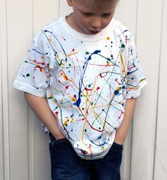 a young boy standing in front of a white wall wearing jeans and a t - shirt with paint splatters on it