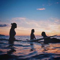 three people are in the water with surfboards at sunset or dawn, and one person is sitting on a surfboard