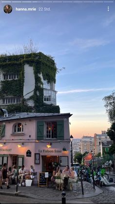 people are sitting outside on the sidewalk in front of a building with plants growing up it's side