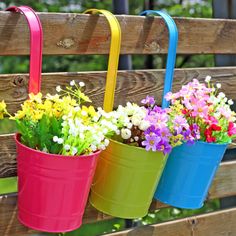 three buckets filled with colorful flowers hanging from a wooden bench in front of trees