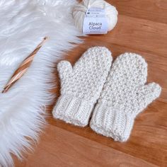 two white knitted mittens sitting on top of a wooden floor next to a ball of yarn