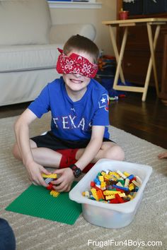 a young boy sitting on the floor next to a bowl of legos with a blind folded over his eyes