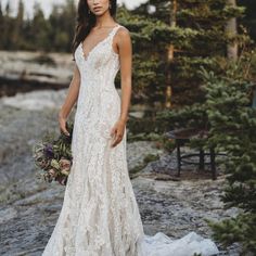 a woman in a white wedding dress standing on rocks near trees and holding a bouquet