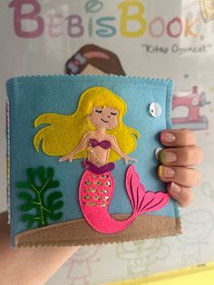 a hand holding up a felt book with a mermaid on it