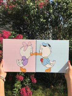 someone holding up two cartoon paintings in front of some bushes and trees with pink flowers