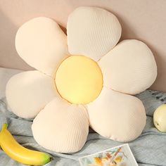 a white flower pillow sitting on top of a bed next to bananas and an apple