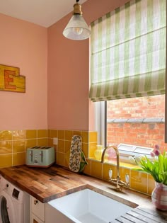 A photo of a kitchen. At the bottom is a dark wood countertop with a white sink in the middle of the counter with gold taps. On the left of the counter is a mint green toaster and a small white and blue tray. The bottom quarter of the wall above the counter is yellow mustard tiles & the rest of the walls are light pink. The ceiling is white with a small cone-shaped glass lampshade. Behind the sink is a window with a white and green vertical striped roman blind half lowered. Eclectic Homes, Verdant Green, Roman Blind, Yellow Walls, Tiny Apartment, Home Upgrades, Bold Stripes, Eclectic Home, Pretty House