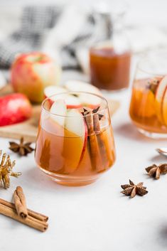 two glasses filled with apple cider next to cinnamon sticks and apples on a table