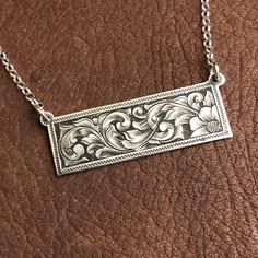 a silver necklace with an ornate design on the front and back of it, sitting on a brown leather surface