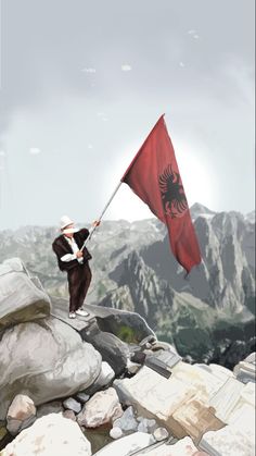 a man holding a red flag on top of a rocky mountain with mountains in the background