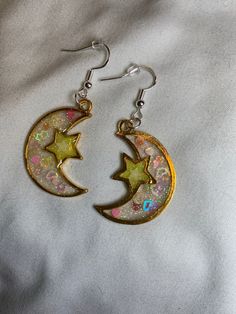 Shine like the night sky with these celestial-themed earrings. The moon and star are meticulously crafted in gold bezel settings. The moon is filled with white and clear glitter UV resin, further adorned with a heart and star-shaped confetti glitter that adds a playful touch. On the other hand, the star boasts neon yellow glitter and a glow-in-the-dark star, casting a unique glow that will certainly turn heads. Magical Gold Jewelry For Party, Celestial Star-shaped Sparkling Earrings, Sparkling Star-shaped Celestial Earrings, Gold Jewelry With Moon Charm For Party, White Moon Charm Jewelry For Party, White Moon Charm Party Jewelry, White Party Jewelry With Moon Charm, Celestial Star Charm Round Earrings, Space-themed Gold Jewelry Gift