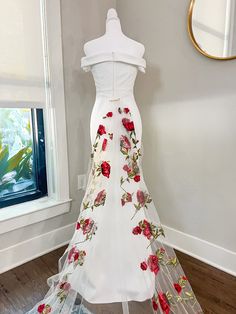 a white dress with red flowers on it is displayed in front of a mirror and window