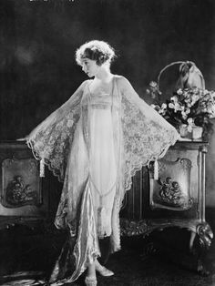 an old fashion photo of a woman wearing a long dress and shawl, standing in front of a dresser with flowers on it