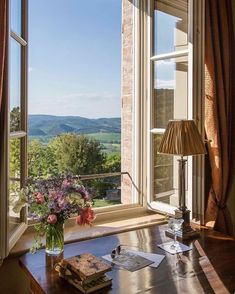 a desk with a book and flowers on it in front of a window overlooking the countryside