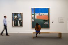 a woman sitting on a bench in an art gallery looking at paintings hanging on the wall