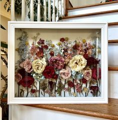 an arrangement of dried flowers in a white frame on the stairs with railings behind it