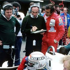 two men in red racing suits and headphones are talking to another man who is writing something on a clipboard