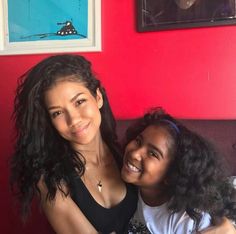 two young women sitting next to each other in front of a red wall and smiling at the camera