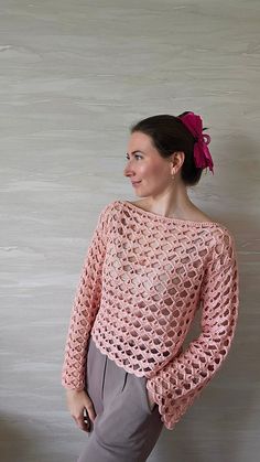 a woman standing in front of a wall wearing a pink crochet top and grey pants