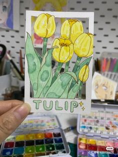 a person holding up a card with yellow tulips in the background and watercolor paints