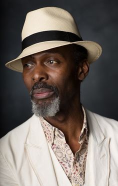 a man wearing a white suit and hat posing for a photo in front of a dark background